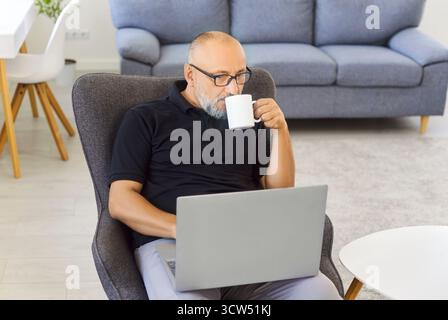 Senior man with laptop drinking coffee while working remotely in cozy home Stock Photo