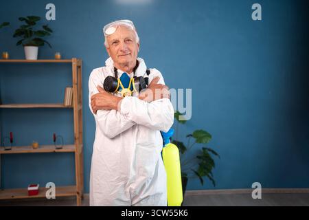 Elderly pest control worker hugging himself and smiling, feeling comfortable inside infested house. Exterminator in protective suit holding spray Stock Photo