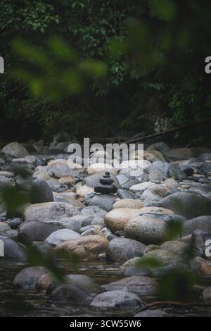 Smooth, rounded stones are carefully stacked on top of each other along the banks of a river, surrounded by lush green foliage and the calming sounds Stock Photo