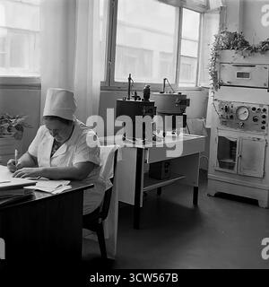 A female laboratory technician works at her desk in the quality control lab of the Slavyansk Bread Factory in this authentic photo from 1978. Dressed in a white uniform, the specialist is focused on her paperwork, surrounded by vintage Soviet scientific equipment like drying ovens and dough testing apparatus. This black and white image documents the crucial, behind-the-scenes role of science and standardization in the USSR's food industry, highlighting the work of skilled female professionals Stock Photo