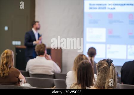 Speaker in dark suit presents to seated audience in modern conference room, large projection screen shows slides, attendees listen attentively during business presentation Stock Photo