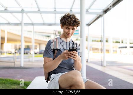 Young man sitting at airport bench using smartphone, focused on screen, Stock Photo
