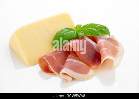Close-up of thinly sliced cured ham served with a block of cheese and fresh basil leaves on a white background, perfect for food photography and culin Stock Photo