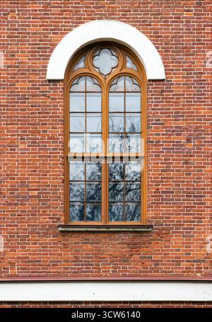 A close-up vertical photo of a weathered red brick exterior featuring a wooden-framed window with Gothic details and white arch. Front view, backgroun Stock Photo