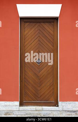 A tall wooden door featuring a chevron pattern sits within a vivid red wall framed by white trim. A small diamond window adds character. Front view, b Stock Photo