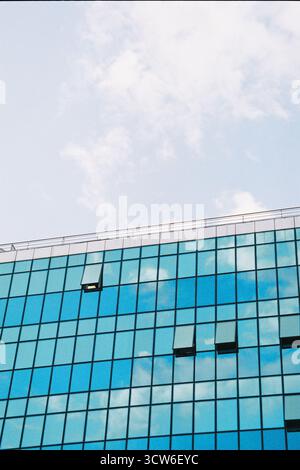 Vertical shot: half glass building with open windows, half bright sky with clouds. Sunlit contrast between structure and nature. Film-like. Stock Photo