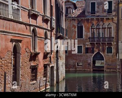 Aged Venetian Palazzi Corner on a Small Canal - A sunlit corner view of historic, decaying Venetian palazzi facades and brickwork, meeting at a small, Stock Photo