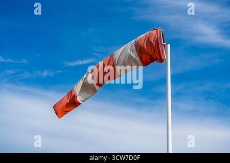 Landscape view of colorful white and orange red windsock aka wind cone or wind sleeve blowing in the air against bright blue sky with wispy clouds Stock Photo