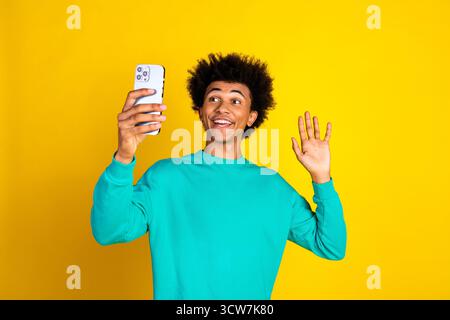 Young man with curly hair smiling and gesturing while taking a selfie on a smartphone against a vibrant yellow background Stock Photo