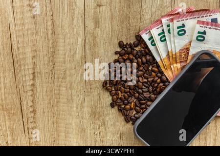Top view of roasted coffee beans, euro banknotes, and smartphone on wooden table. Symbol of rising coffee prices, inflation, global economy, and moder Stock Photo