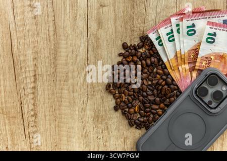 Top view of roasted coffee beans, euro banknotes, and smartphone on wooden table. Symbol of rising coffee prices, inflation, global economy, and moder Stock Photo