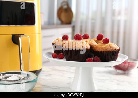 Freshly baked muffins with raspberries and air fryer on white marble table in kitchen, closeup Stock Photo