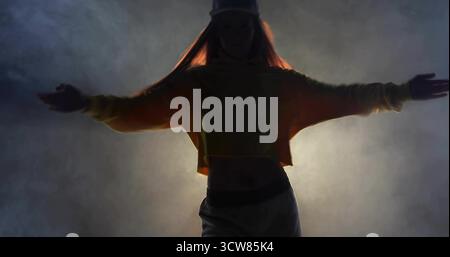 Striking dancer holding arms out on stage, with cap cropped hoodie sweatpants spotlight fog effect. Performance, art, expression, stage, lighting, atm Stock Photo