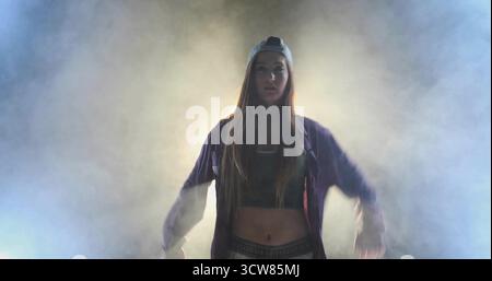 Performing female dancer wearing backwards cap and zip-up jacket, holding arms out on foggy stage. Contemporary, performance, theatrical, moody, mysti Stock Photo