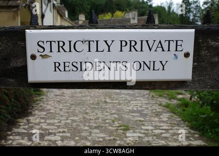 Sign reading 'Strictly Private Residents Only' mounted on a wooden gate. The background features a cobblestone path leading towards stone buildings an Stock Photo