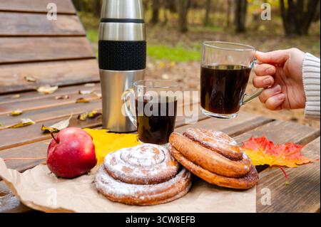 Cozy autumn picnic scene with stainless steel thermos, two glass cups of coffee, sweet cinnamon rolls with powdered sugar, red apple and colorful mapl Stock Photo