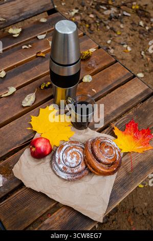Warm autumn still life with a stainless steel thermos, two glass mugs of coffee, sweet cinnamon rolls, a red apple, and colorful maple leaves arranged Stock Photo