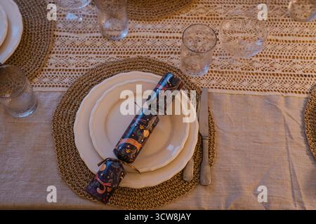 Elegant Christmas table setting with white plates, woven placemats, crystal glasses and festive crackers on a linen tablecloth with crochet details. Stock Photo