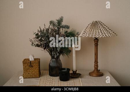 Cozy winter home decor with a bouquet of pine branches, cones, eucalyptus in a glass vase, vintage wooden lamp and woven tissue box on a linen-covered Stock Photo