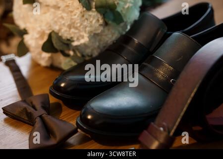 Close-up showcasing groom's black leather loafers, matching bowtie, and a white floral bouquet. luxury wedding accessories on wooden table, romantic m Stock Photo