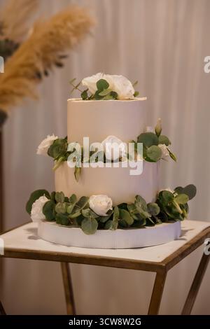 A beautifully decorated two-tier wedding cake stands on a wooden stand. adorned with white roses and eucalyptus greenery, this dessert symbolizes love Stock Photo
