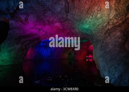 The interior of a quarry cave with dark water and rock walls illuminated by a dynamic spectrum of colorful light, including pink, blue, and green Stock Photo