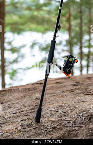 Close-up of a spinning rod with a lure, set against a blurred lake and tree background, ready for fishing. Stock Photo