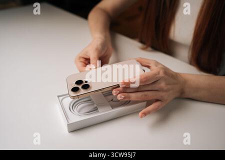 High-angle view of woman unpacking new iPhone 16 pro from white box on table, showcasing latest apple technology in modern setting Stock Photo