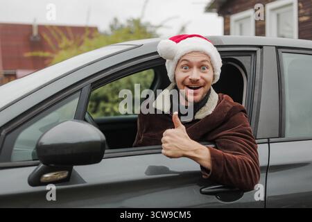 Young happy man in Christmas Santa hat showing thumbs up while driving car. Stock Photo