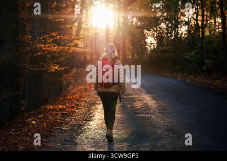 Woman with backpack walking on road in woodland during sunset. Female tourist hiking in autumn forest Stock Photo