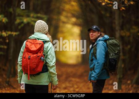 Two hikers in sports outdoor clothing with backpacks exploring forest. Hiking couple walks on trekking trail in autumn woodland together Stock Photo