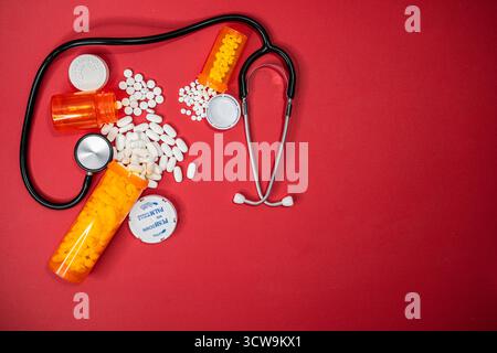 Three RX containers spilled out on a table top surrounded by a black Stethoscope. White pills intermingled on a red background. Stock Photo