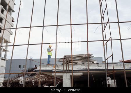 Caucasian middle aged man wearing safety helmet standing on construction site using smartphone, surrounded by steel framework and building materials during workday Stock Photo