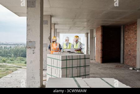 Three builders wearing safety helmets and vests, standing at construction site discussing building plans on unfinished concrete floor Stock Photo