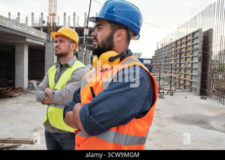 Two builders standing on construction site wearing safety helmets and vests, crossing arms and observing building progress together Stock Photo