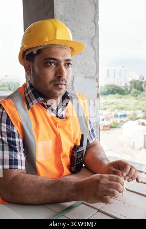 Male builder wearing construction helmet and safety vest standing at building site holding toothpick looking away with serious expression Stock Photo