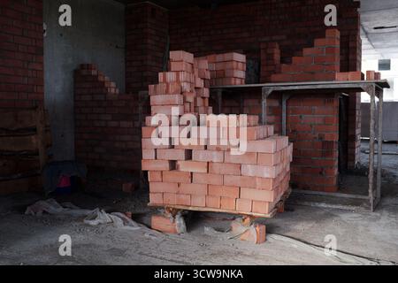 Stack of red bricks arranged on wooden pallet standing inside unfinished building under construction, exposed brick walls and concrete floor visible in background, no people present Stock Photo