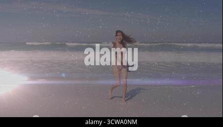 Running adult woman wearing floral bikini holding towel on sandy beach, sun flare and ocean waves Stock Photo