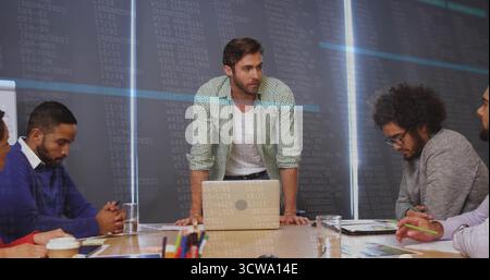 Standing project manager leaning forward at meeting table in office with laptop and data display Stock Photo