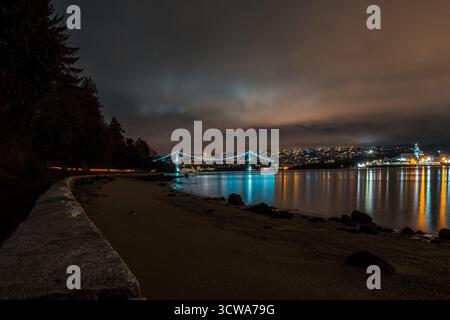 A serene waterfront scene shows city lights reflecting on the calm water at night. Soft clouds drift above, and a bridge connects the two shores. Stock Photo