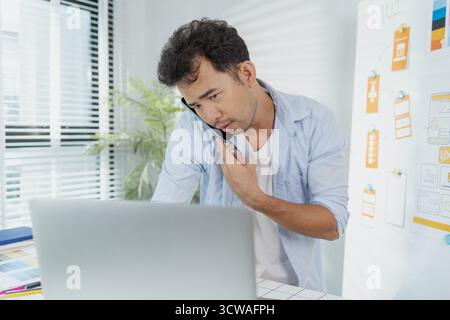 A creative designer works on UX/UI planning, analyzing sketches and prototypes on a whiteboard while using a laptop. The process shows design thinking Stock Photo