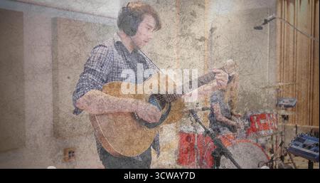 Strumming guitarist playing acoustic guitar in soundproof studio, with drummer behind red drum kit Stock Photo