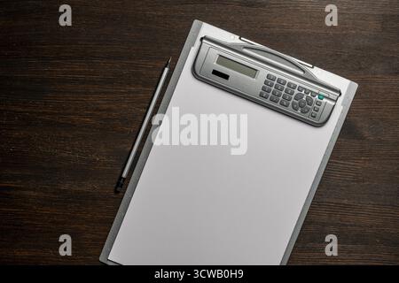 Clipboard with a white blank paper and pencil on wooden table, close up, top view. Grey desktop with electronic calculator and a ruler Stock Photo