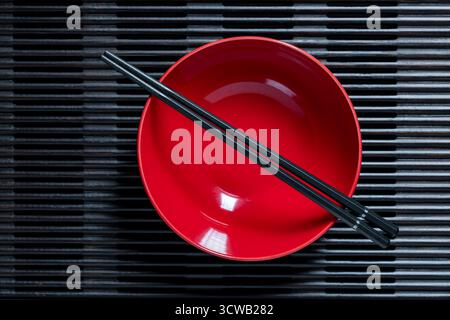 A pair of black chopsticks lying across an empty red soup bowl on wooden striped tray, close up, top view Stock Photo