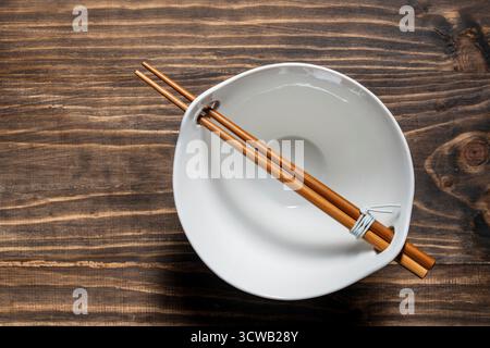 A pair of chopsticks lying across an empty white soup bowl on wooden table, close up, top view Stock Photo