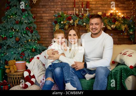 Happy family of three sitting on a sofa in matching white sweaters, smiling at the camera in a warmly decorated Christmas room Stock Photo