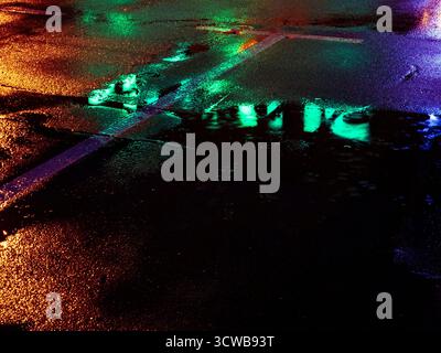 Wet urban asphalt reflecting bright neon lights at night, creating an abstract and colorful scene with dynamic patterns and glowing puddles, conveying Stock Photo