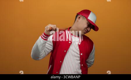 Young man in a baseball uniform poses confidently with a bat against an orange background, showcasing a sporty and vibrant atmosphere. Stock Photo