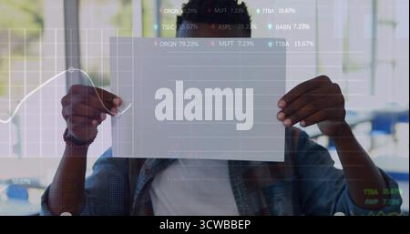 Holding man wearing denim jacket obscuring face with paper in office cafe with line chart overlay Stock Photo