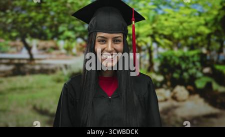 Young hispanic woman in graduation gown smiling joyfully in an outdoor park setting, celebrating academic achievement under sunny skies. Stock Photo